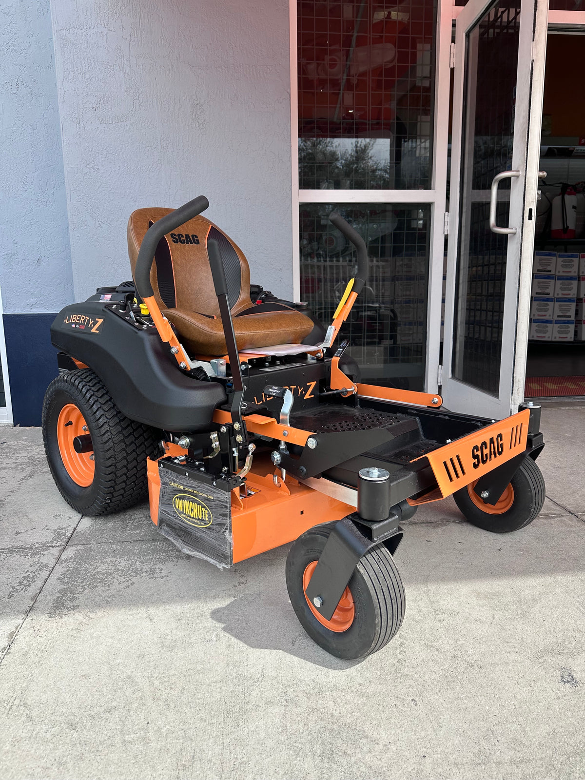 An orange and black zero-till riding lawn mower with a brown seat, parked outdoors on a concrete surface in front of a building with glass doors. The mower has 'SCAG' branding on the seat, deck, and front bumper.